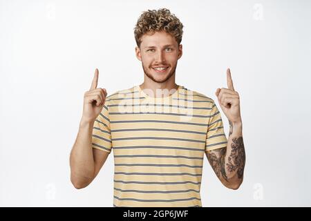 Handsome blond man smiles and shows advertisement on top, pointing fingers up at logo or company sign, standing over white background Stock Photo