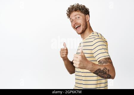 Handsome hipster guy shows thumbs up and smiles happy, congratulating with great work, praising you, standing satisfied against white background Stock Photo