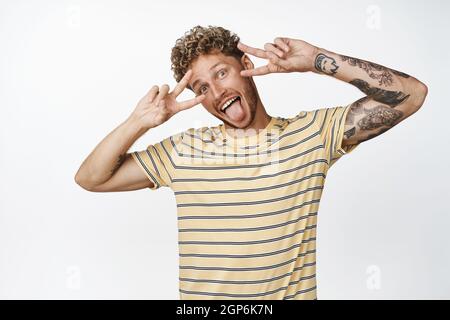 Funny blond man shows tongue and v-signs peace near face, making funny faces and standing over white background in casual clothes Stock Photo