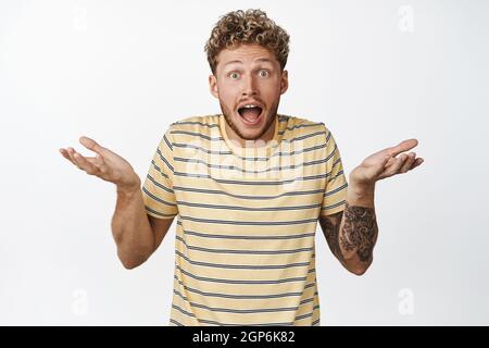 Excited young man looking disbelief, shrugging shoulders and staring surprised at camera, cant believe he won prize, standing confused against white Stock Photo