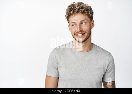Portrait of attractive blond guy smiling, looking aside at copy space, wearing grey t-shirt, standing against white background Stock Photo