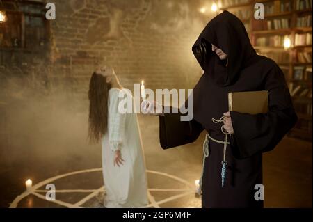 Male exorcist in black hood casting out demons from a woman. Exorcism, mystery paranormal ritual, dark religion, night horror Stock Photo