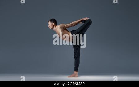 Male yoga keeps balanc in a difficult pose, grey background. Strong man doing yogi exercise, asana training, top concentration, healthy lifestyle Stock Photo