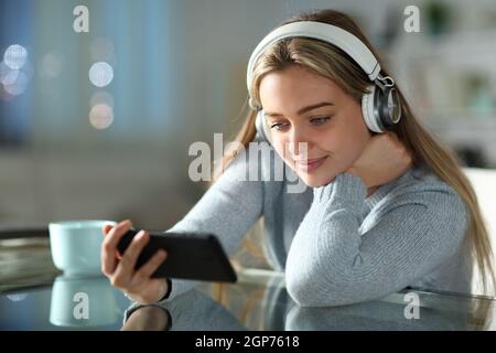 Teen watching videos on smart phone wearing headphones in the night in the living room at home Stock Photo picture