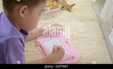 Happy Asian klid little boy preschool writing at the magnetic drawing board at home on carpet. Funny child playing magnetic drawing board. Education L Stock Photo