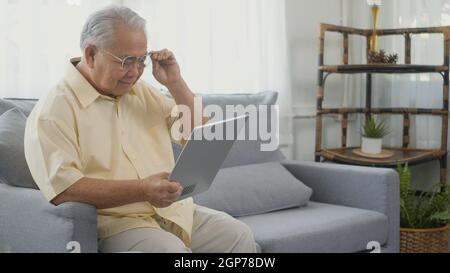 Senior man dressed to wear eyeglasses sitting on sofa looking news on digital tablet computer in living room at home, Happy old man retired using digi Stock Photo