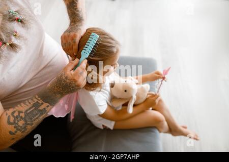 Man with scrunchies in beard brushes girl hair in living room Stock Photo