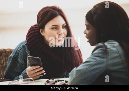 Two young beautiful girls sit at a table talking and using the mobile phone. Stock Photo