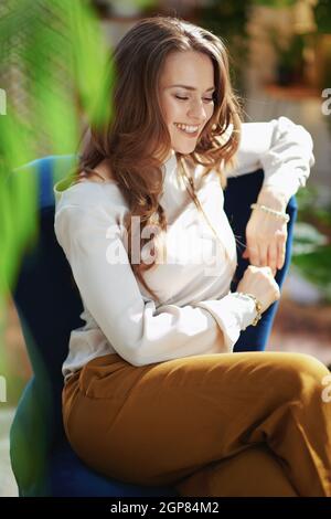 Green Home. smiling stylish middle aged housewife with long wavy hair in the modern living room in sunny day in green pants and grey blouse sitting in Stock Photo