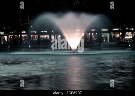 A long exposure shot of a mountain lake fountain spurting water with shops in the background Stock Photo