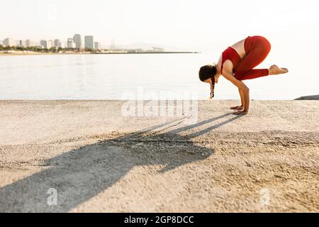 Young hispanic woman practicing yoga and doing bakasana position by the sea Stock Photo