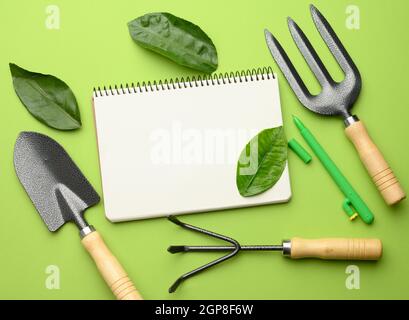 open notebook with blank white sheets and various gardening tools with wooden handles on green background, flat lay, copy space Stock Photo