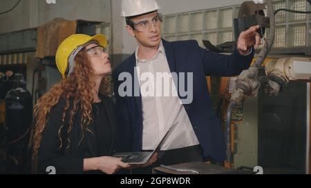 Caucasian maintenance engineer man and diverse female factory workers in hardhats talking and present or explain robot welding machine on plant, Manag Stock Photo