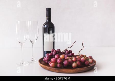 Red wine bottle, large burgundy fresh grapes on wooden dish, empty wine glasses on white background, mockup. Stock Photo