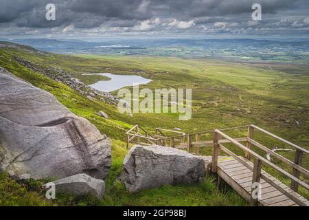 Massive boulders and wooden stairs of Cuilcagh Park boardwalk leading down to the valley with lake from Cuilcagh Mountain peak, Northern Ireland Stock Photo