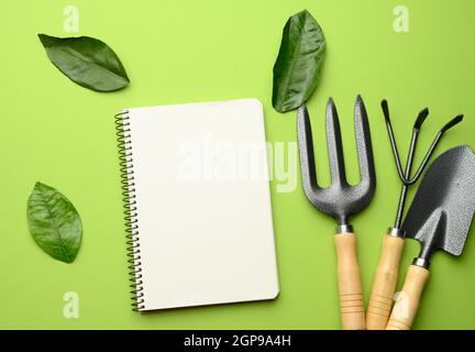 open notebook with blank white sheets and various gardening tools with wooden handles on green background, flat lay, copy space Stock Photo