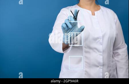 woman doctor in a white medical coat, blue latex gloves stands on a blue background, hand touches and notes in the list on a virtual screen Stock Photo