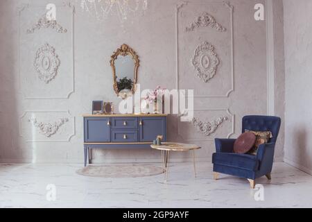 Luxurious interior with a chest of drawers and velvet armchair in a classic style, marble floor, stucco decor on the walls Stock Photo