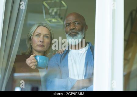 Worried senior diverse couple looking through window and embracing Stock Photo