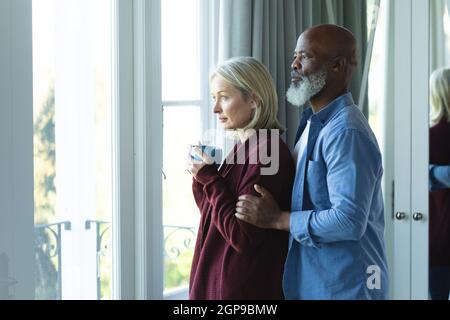 Worried senior diverse couple looking through window and embracing Stock Photo