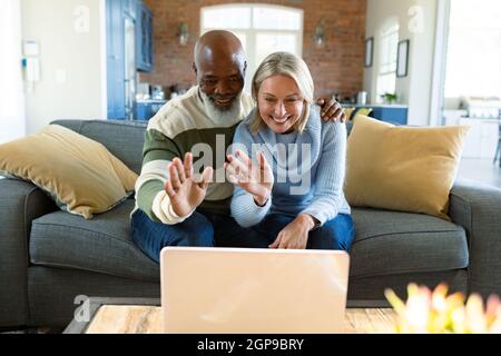Happy senior diverse couple in living room sitting on sofa, using laptop, making video call Stock Photo