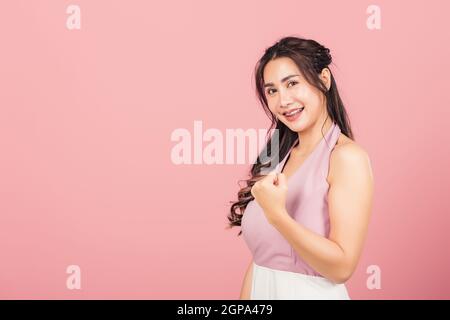 Happy Asian portrait beautiful cute young woman stand makes raised fists up celebrating her winning success gesture, studio shot isolated pink backgro Stock Photo