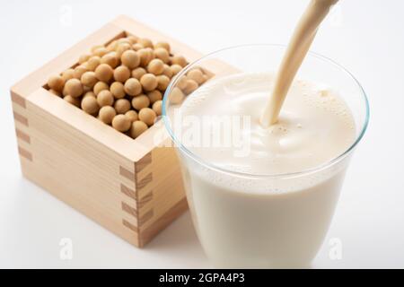 Pour soy milk into a glass placed on a white background. Soybeans in a Masu in the background Stock Photo