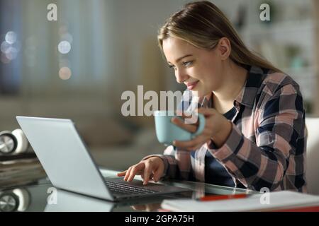 Satisfied student e-learning using laptop holding coffee cup at home in the night Stock Photo