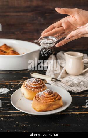 Woman sprinkling powdered sugar onto tasty cinnamon rolls on table Stock Photo