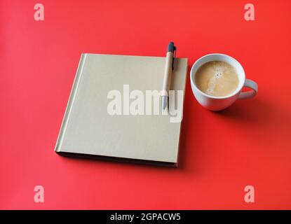 Photo of closed blank square book, coffee cup and pen on red paper background. Template for placing your design. Stock Photo