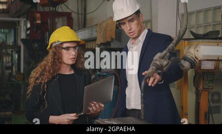 Caucasian maintenance engineer man and diverse female factory workers in hardhats talking and present or explain robot welding machine on plant, Manag Stock Photo