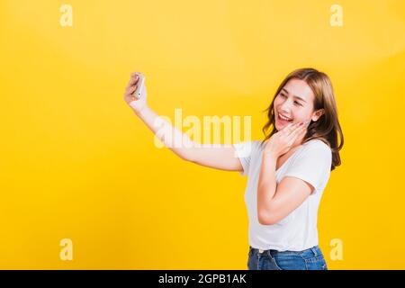 Asian Thai portrait happy beautiful cute young woman smiling wear t-shirt making selfie photo or video call on smartphone looking the phone, studio sh Stock Photo