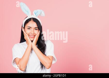 Happy Easter Day. Beautiful young woman teen smiling wearing Easter rabbit bunny ears holding her cheeks excited surprised, Portrait female face touch Stock Photo