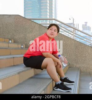 Overweight woman with a bottle of fresh drinking water resting on steps in the city after work out Stock Photo