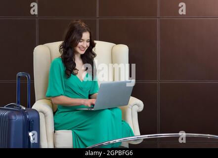 Young Brazilian tourist in green clothes typing on laptop computer keyboard at the Business Lounge Airport service. Her luggage placed beside the armc Stock Photo