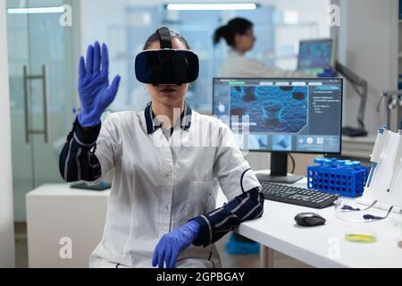Biologist researcher wearing virtual reality headset examining biochemistry experiment working in microbiology hospital laboratory using biotechnology. Specialist scientist analyzing virus development Stock Photo