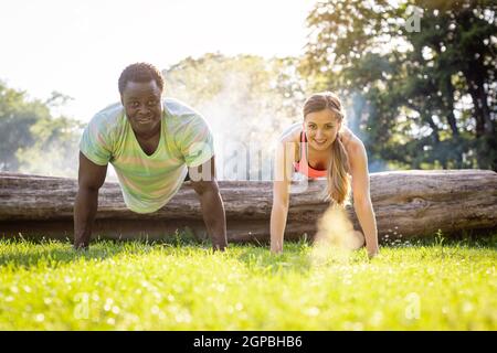 Woman and her fitness coach doing push-ups on tree trunk in summer looking at each other Stock Photo