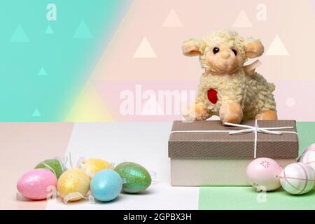 Happy Easter greeting card. Decorative composition of colorful eggs, a gift box with a easter lamb on it on a multicolour table in front of modern abs Stock Photo