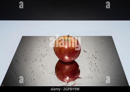 A red apple resting on a mirror with falling water drops Stock Photo