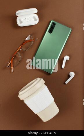 Smartphone, reusable coffee cup, wireless earphones and glasses on brown background top view Stock Photo