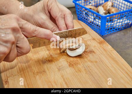 Woman cuts mushrooms on wooden cutting board. Cooks according to the recipe at home in the kitchen. Stock Photo
