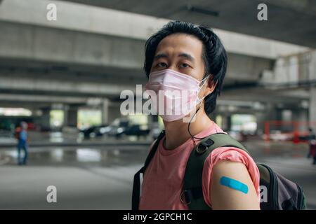 Young Asian man wearing covid-19 face protection mask looking at bandage after vaccine for protection against corona virus during day wearing a bag Stock Photo