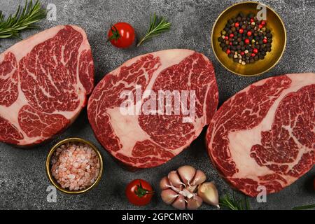 Close up three aged prime marbled raw ribeye beef steaks with spices, over stone table background, elevated top view, directly above Stock Photo