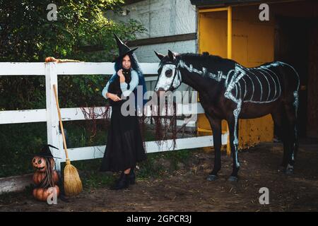 A girl dressed as a witch on a halloween party holds a black cat in her arms and stands by a corral on a farm next to a horse