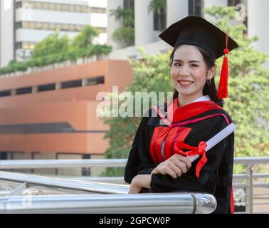 Portrait of happy graduated woman in black graduation gowns holding diploma and standing with arms crossed. Smiling student wearing mortarboard cap lo Stock Photo