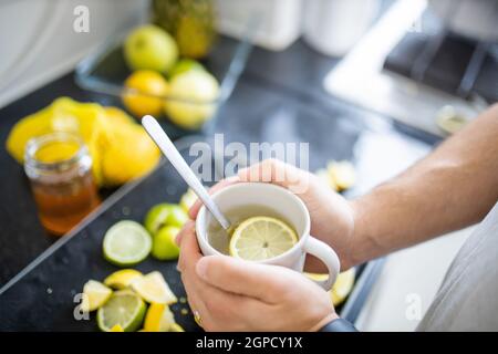 Male hands holding cup of lemon tea above black tray with slices of limes and lemons. Man with cup of tea with lemon slice floating inside. Sweet citr Stock Photo