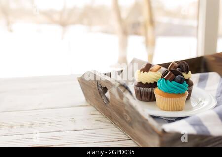 Tasty multicolored muffins and cupcakes on the white plate on a wooden tray with towel. Decorated with different candies, cookies and colored sweet cr Stock Photo