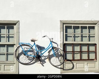 A white house wall with two windows with white wood decorations. A white and blue bicycle hangs on the wall between the two windows. Stock Photo