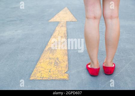 Girl wear red shoes  walking towards with yellow traffic arrow signage on an asphalt road background Stock Photo