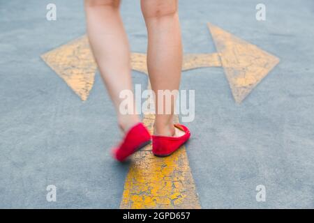 Girl wear red shoes  walking towards with yellow traffic arrow signage on an asphalt road background Stock Photo